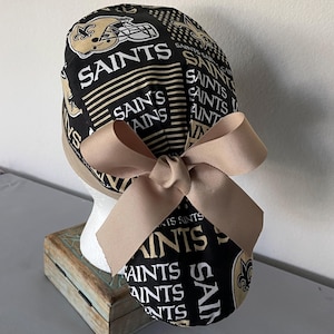 Op de afbeelding: Een zwarte operatiemuts met een herhalend patroon van het New Orleans Saints-logo, footballhelmen en het woord SAINTS in witte en gouden letters. De muts is voorzien van een effen beige lint dat aan de achterkant tot een strik is geknoopt.