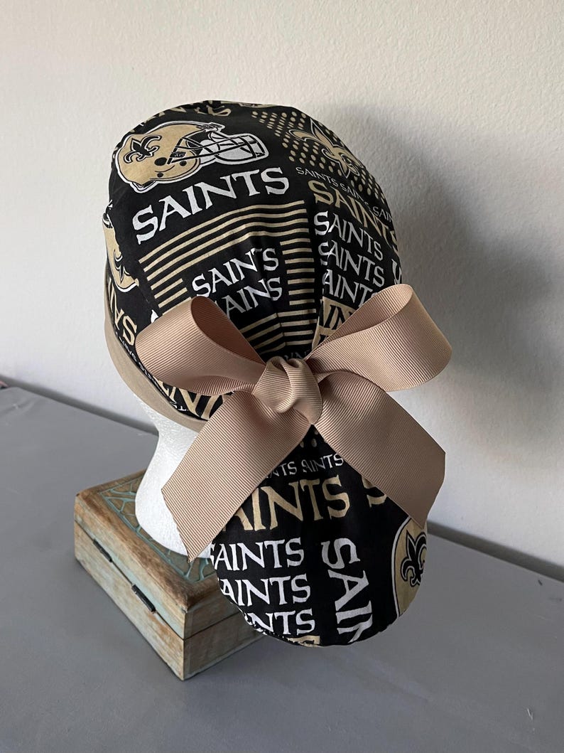 Op de afbeelding: Een zwarte operatiemuts met een herhalend patroon van het New Orleans Saints-logo, footballhelmen en het woord SAINTS in witte en gouden letters. De muts is voorzien van een effen beige lint dat aan de achterkant tot een strik is geknoopt.