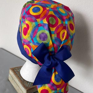 May include: A colorful surgical scrub hat with a blue bow. The hat features a pattern of overlapping circles in shades of yellow, orange, red, green, blue, and purple.