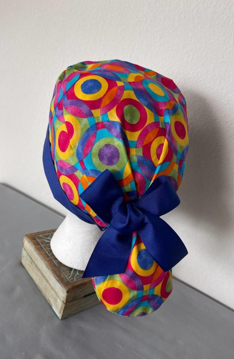 May include: A colorful surgical scrub hat with a blue bow. The hat features a pattern of overlapping circles in shades of yellow, orange, red, green, blue, and purple.