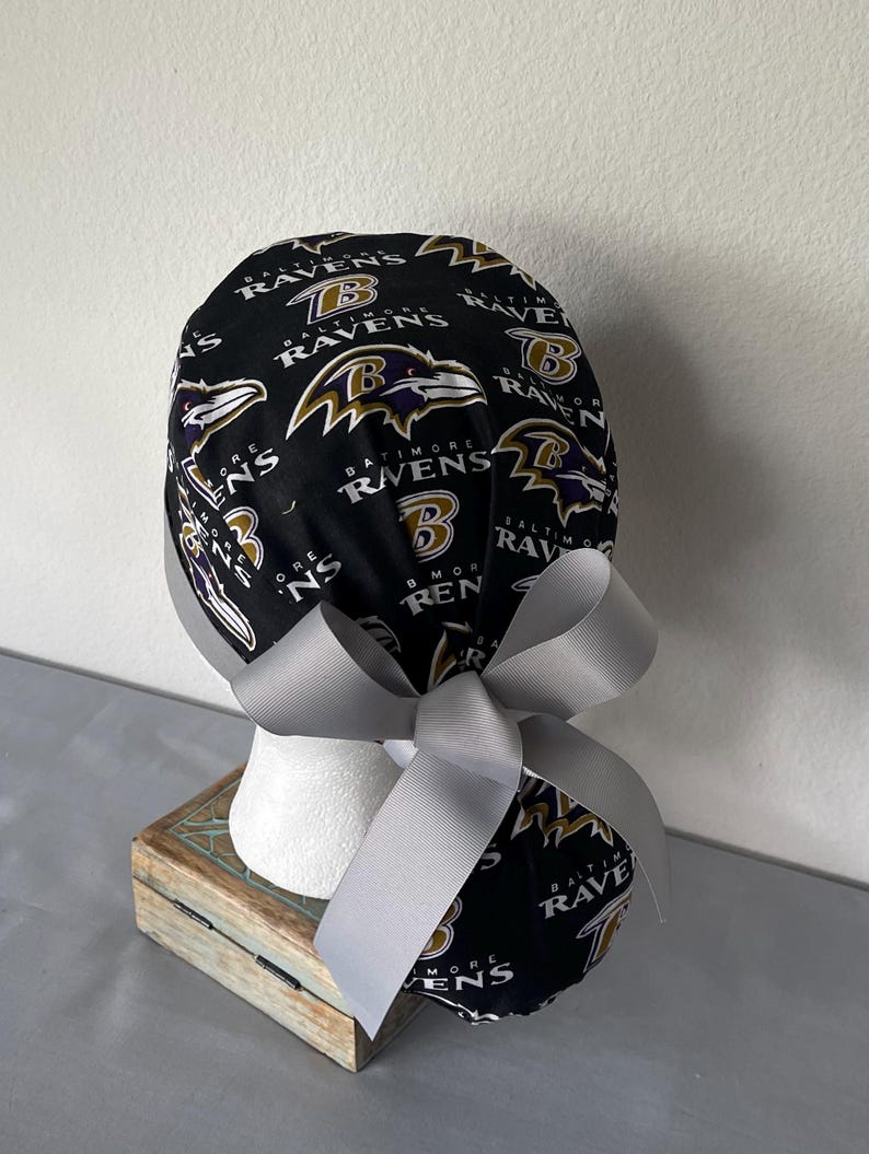 Op de afbeelding: Een zwarte operatiemuts met een herhalend patroon van het Baltimore Ravens logo en tekst in paars, goud en wit. De muts is voorzien van een effen lichtgrijs lint dat aan de achterkant tot een strik is geknoopt voor een verstelbare pasvorm.