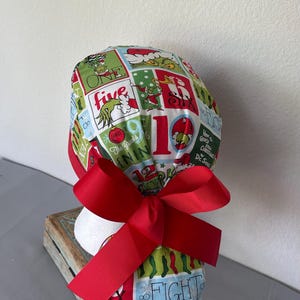 Op de afbeelding: Een operatiemuts met een kleurrijk ruitpatroon met illustraties van de Grinch, kerstsokken en diverse cijfers. De stof is overwegend groen, rood en wit en de muts wordt aan de achterkant vastgemaakt met een breed, effen rood lint.