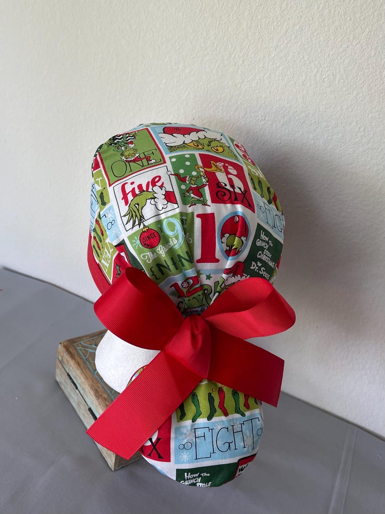 Op de afbeelding: Een operatiemuts met een kleurrijk ruitpatroon met illustraties van de Grinch, kerstsokken en diverse cijfers. De stof is overwegend groen, rood en wit en de muts wordt aan de achterkant vastgemaakt met een breed, effen rood lint.