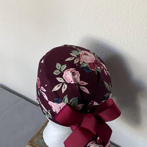 May include: A burgundy scrub cap featuring a floral pattern with light pink roses and green leaves. The cap includes a matching solid burgundy ribbon tied into a bow at the back, designed to secure the fabric in a gathered style around the head.
