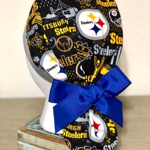 Könnte beinhalten: Schwarze und gelbe chirurgische Mütze im Pittsburgh Steelers-Design mit einer blauen Schleife. Die Mütze zeigt verschiedene Steelers-Logos und Grafiken, darunter das Teamlogo, die Wörter "Steelers" und "Pittsburgh" sowie ein Hotdog.