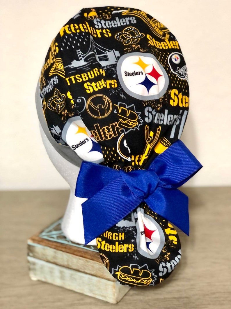 Op de afbeelding: Een zwarte operatiemuts met een herhalend Pittsburgh Steelers-patroon, gele en witte tekst, teamlogo's en stadsafbeeldingen. De muts is voorzien van een grote, effen blauwe strik aan de achterkant, vastgezet met een geplooid ontwerp van de stof.