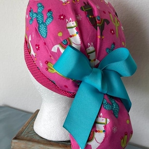May include: Pink surgical scrub hat with a turquoise bow. The hat has a pattern of llamas and cacti.