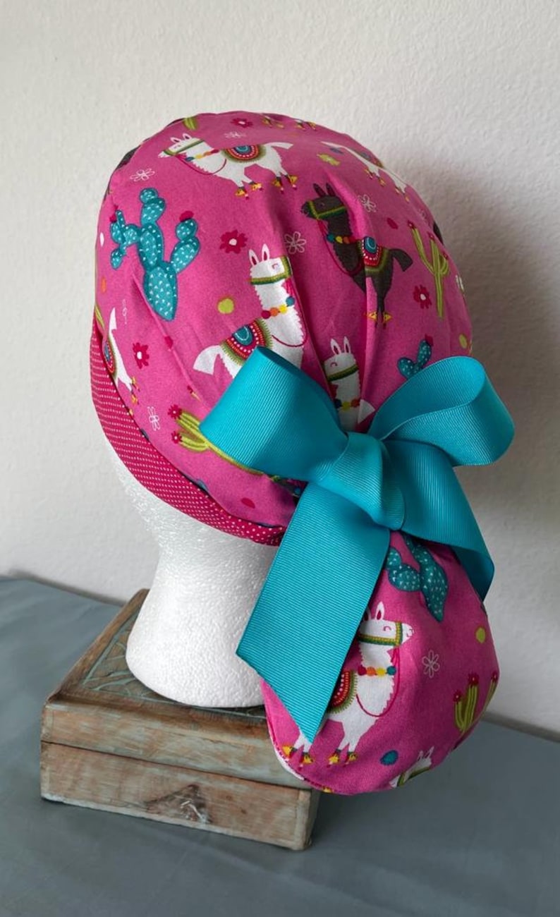May include: Pink surgical scrub hat with a turquoise bow. The hat has a pattern of llamas and cacti.