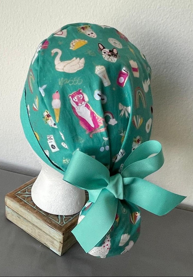May include: Teal blue surgical scrub hat with a colorful pattern of animals, rainbows, and other whimsical designs. The hat has a matching teal blue ribbon tied in a bow at the back.