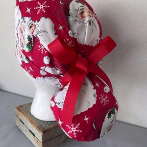 Op de afbeelding: Een rode operatiemuts met een herhalend patroon van kerstmangezichten, witte sneeuwvlokken en kleine groene hulstblaadjes. De muts is voorzien van een effen rood lint dat aan de achterkant tot een strik is geknoopt om de stof vast te zetten.