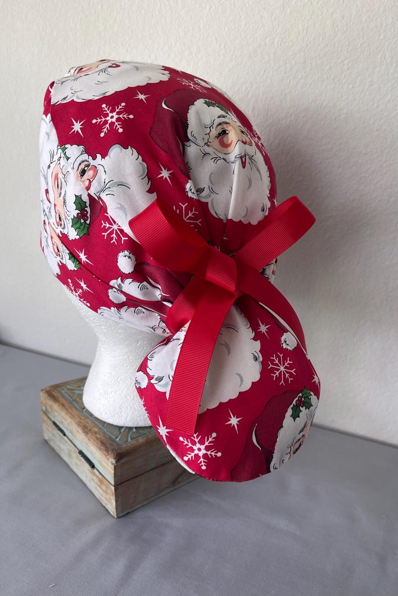 Op de afbeelding: Een rode operatiemuts met een herhalend patroon van kerstmangezichten, witte sneeuwvlokken en kleine groene hulstblaadjes. De muts is voorzien van een effen rood lint dat aan de achterkant tot een strik is geknoopt om de stof vast te zetten.