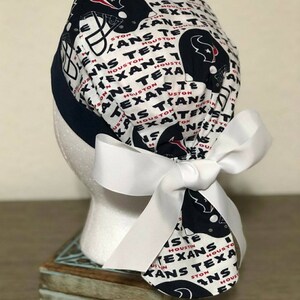 Op de afbeelding: Een operatiemuts met een witte achtergrond en een herhalend patroon van marineblauwe Houston Texans footballhelmen en de tekst Houston Texans in zwart en rood. De muts heeft een effen marineblauwe band en een grote witte strik.