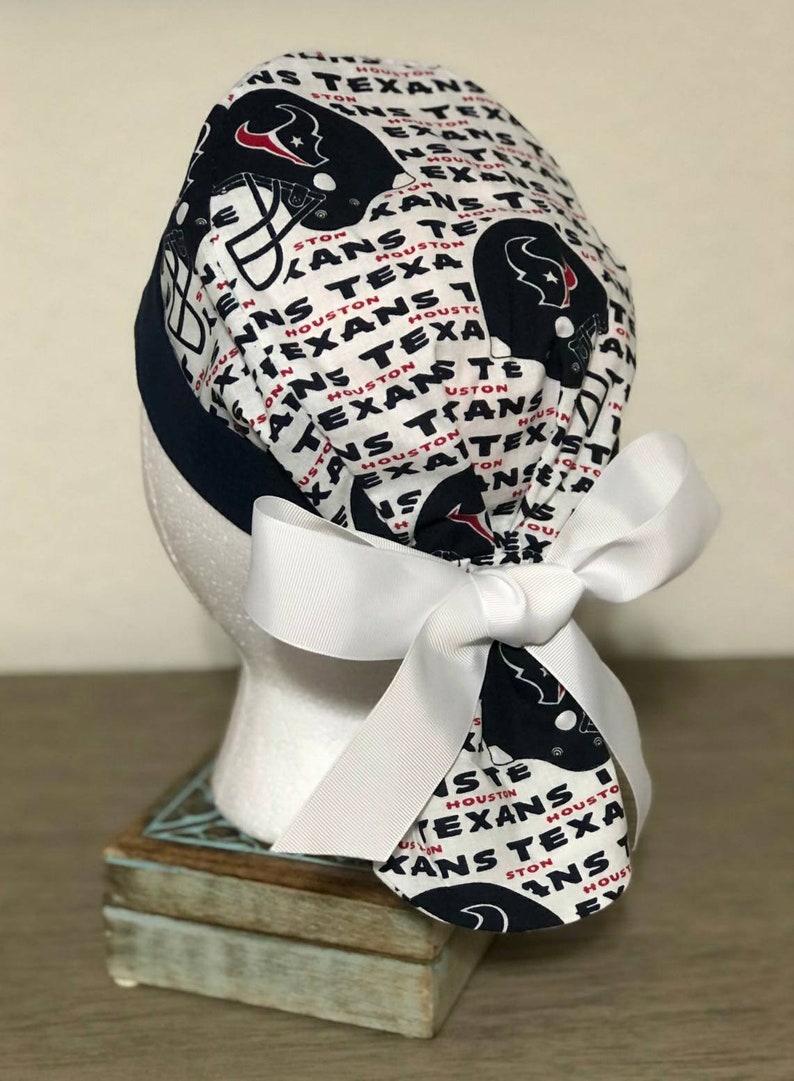 Op de afbeelding: Een operatiemuts met een witte achtergrond en een herhalend patroon van marineblauwe Houston Texans footballhelmen en de tekst Houston Texans in zwart en rood. De muts heeft een effen marineblauwe band en een grote witte strik.