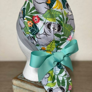 May include: A turquoise scrub hat with a koala and tropical leaf print. The hat has a turquoise ribbon tied in a bow at the back.