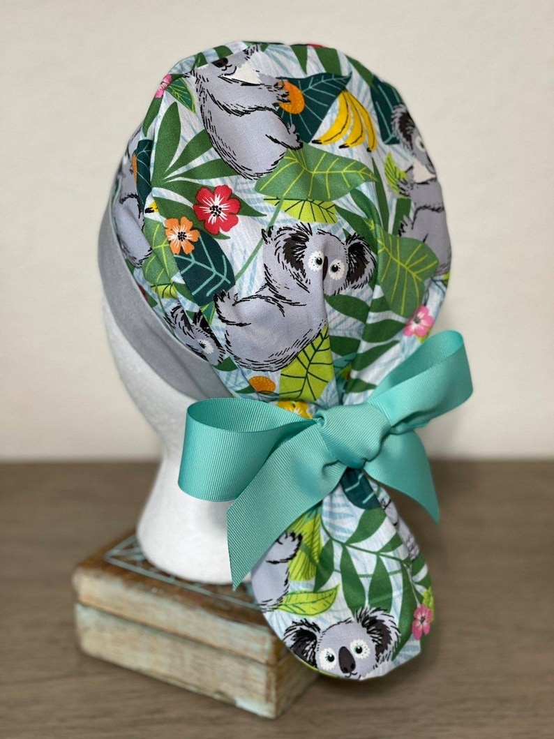 May include: A turquoise scrub hat with a koala and tropical leaf print. The hat has a turquoise ribbon tied in a bow at the back.