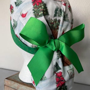 Op de afbeelding: Een witte operatiemuts met een kerstprint van groene dennenbomen, rode kardinalen en de tekst Merry Christmas. De muts is voorzien van een felgroen grosgrain lint dat aan de achterkant is gestrikt tot een grote strik om het model vast te zetten.