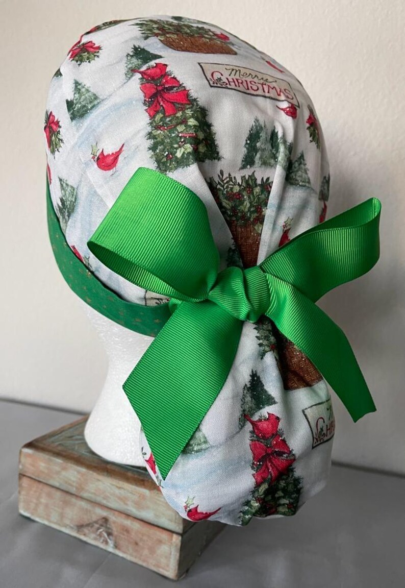Op de afbeelding: Een witte operatiemuts met een kerstprint van groene dennenbomen, rode kardinalen en de tekst Merry Christmas. De muts is voorzien van een felgroen grosgrain lint dat aan de achterkant is gestrikt tot een grote strik om het model vast te zetten.