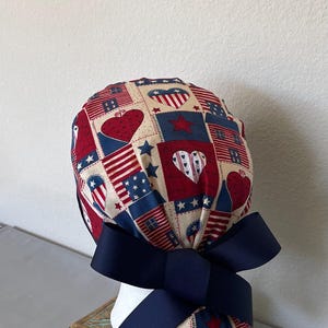 Op de afbeelding: Een operatiemuts met een patchworkpatroon van rode, witte en blauwe vierkanten met hartjes, sterren en strepen. De muts is aan de achterkant vastgezet met een effen marineblauw grosgrain lint dat tot een strik is geknoopt voor een geplooid effect.