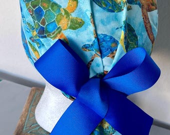 May include: A blue scrub cap featuring a pattern of sea turtles in shades of blue, green, and yellow. A large, vibrant blue bow is attached to the cap. The cap is displayed on a white mannequin head.