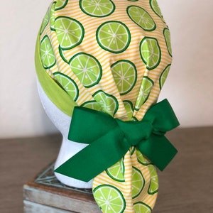 May include: A yellow and green patterned surgical scrub hat with a green bow. The hat has a lime pattern on a yellow and white striped background.