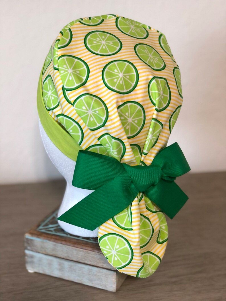 May include: A yellow and green patterned surgical scrub hat with a green bow. The hat has a lime pattern on a yellow and white striped background.
