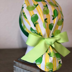 May include: A white and yellow scrub hat with a lime pattern and a green bow. The hat is tied at the back of the head.