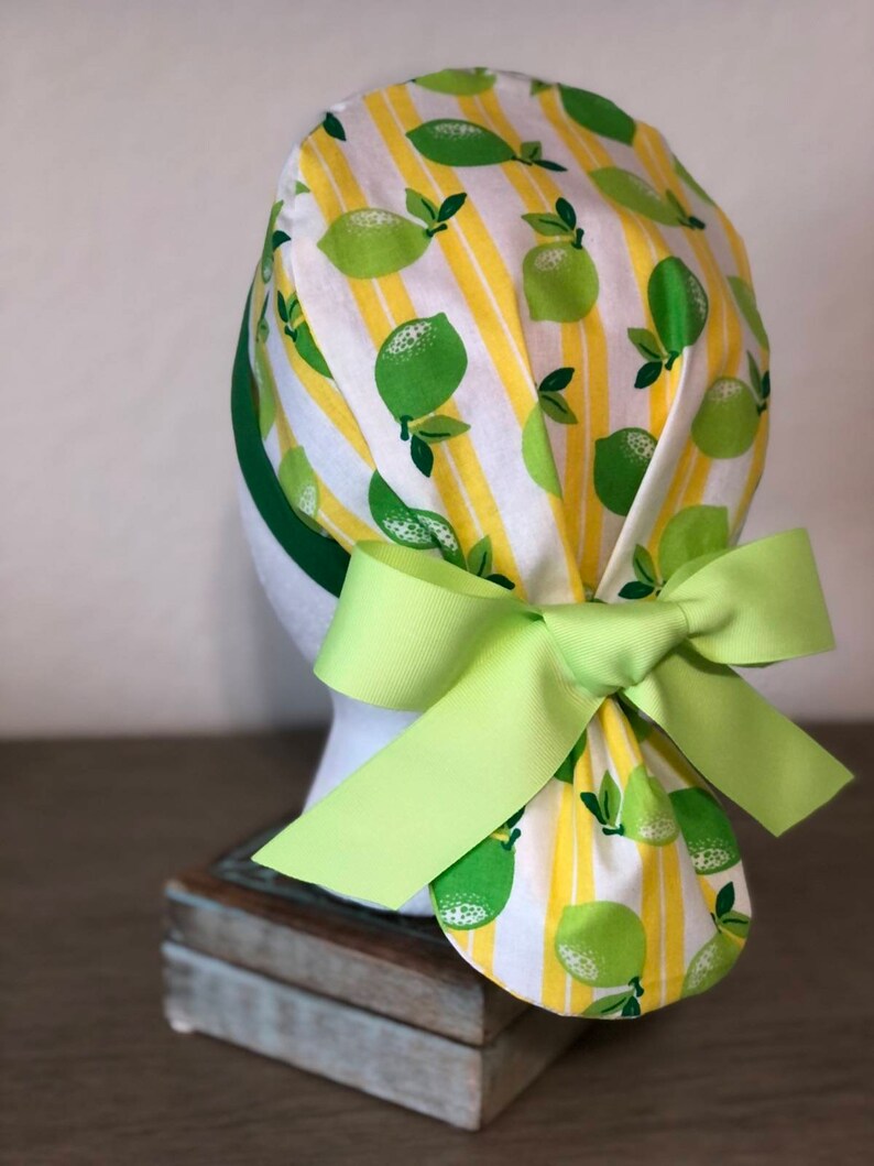 May include: A white and yellow scrub hat with a lime pattern and a green bow. The hat is tied at the back of the head.