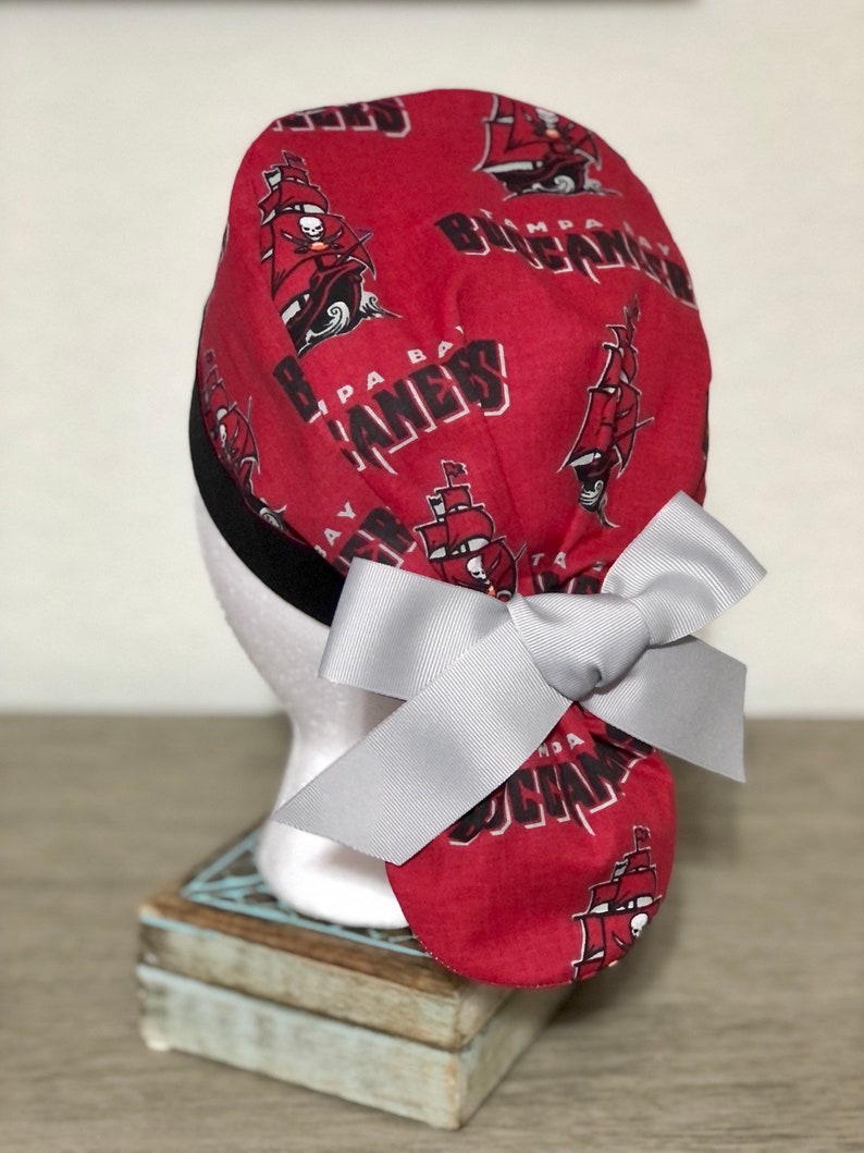 Op de afbeelding: Een rode operatiemuts met een herhalend patroon van Tampa Bay Buccaneers-logo's en piratenschepen, geaccentueerd met een zwarte rand en een grote, lichtgrijze strik aan de achterkant. De stof toont de teamnaam in zwarte en witte letters.