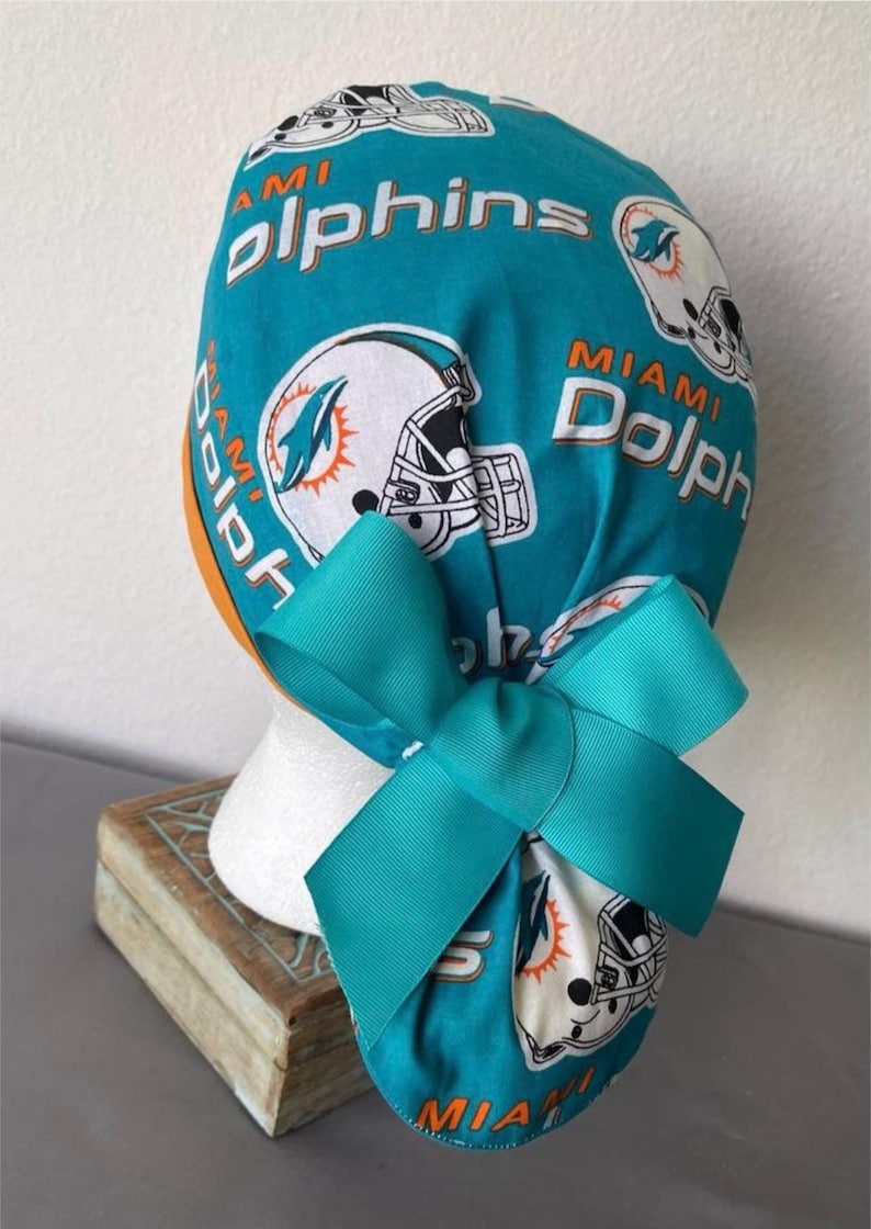 Op de afbeelding: Een turquoise operatiemuts met een herhalend patroon van witte Miami Dolphins footballhelmen en de tekst Miami Dolphins in oranje en wit. De muts is voorzien van een grote, effen turquoise strik van grosgrain lint aan de achterzijde.