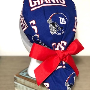 Op de afbeelding: Een blauwe operatiemuts met een herhalend patroon van New York Giants footballhelmen en het woord GIANTS in witte letters met rode randen. De muts is aan de achterkant gestrikt met een brede, effen rode grosgrain lintstrik.