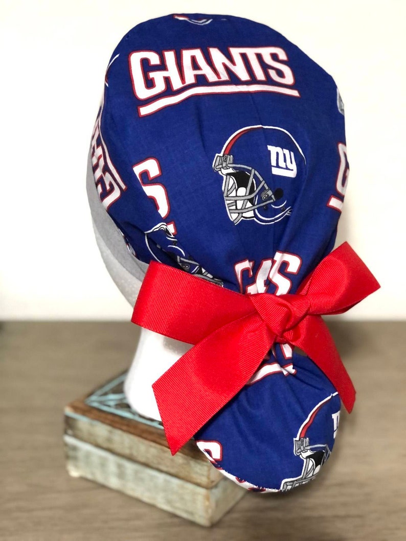 Op de afbeelding: Een blauwe operatiemuts met een herhalend patroon van New York Giants footballhelmen en het woord GIANTS in witte letters met rode randen. De muts is aan de achterkant gestrikt met een brede, effen rode grosgrain lintstrik.