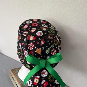 Op de afbeelding: Een zwarte operatiemuts met een feestelijk patroon van koffiebekers, donuts, peperkoekmannetjes en sneeuwvlokken. De muts is voorzien van een felgroen lint dat aan de achterkant tot een strik is gebonden om de stof in een staart te houden.