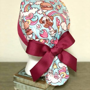 May include: A blue and pink patterned surgical scrub hat with a burgundy bow. The pattern features unicorns, rainbows, pugs, and other whimsical designs.