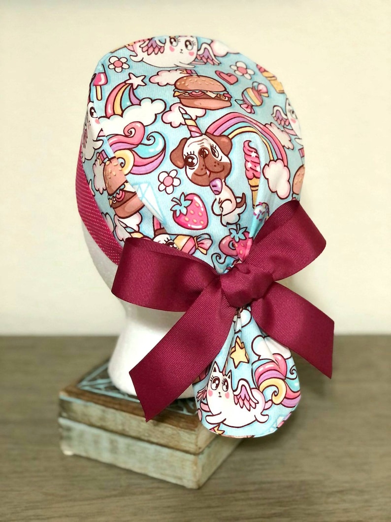 May include: A blue and pink patterned surgical scrub hat with a burgundy bow. The pattern features unicorns, rainbows, pugs, and other whimsical designs.
