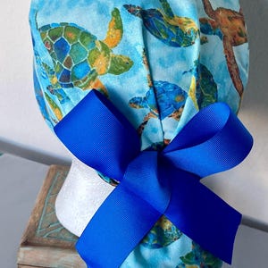 May include: A blue scrub hat with a colorful sea turtle print and a bright blue bow. The hat is made of soft, comfortable fabric and is perfect for nurses, doctors, and other healthcare professionals.