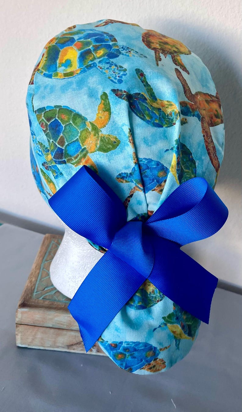 May include: A blue scrub hat with a colorful sea turtle print and a bright blue bow. The hat is made of soft, comfortable fabric and is perfect for nurses, doctors, and other healthcare professionals.