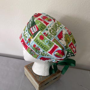 May include: A scrub cap featuring a colorful pattern of The Grinch, Christmas presents, and text including Who Hash, The Grinch, and Nine. The fabric is predominantly green, red, and white, with a dark green ribbon tie at the back for adjustment.