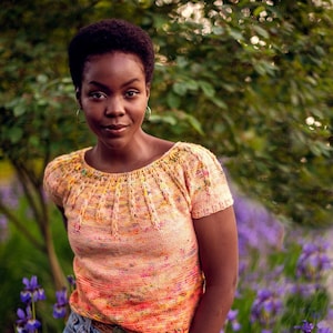 May include: A woman wearing a peach-colored knitted top with a colorful, textured design and a light blue denim skirt. She is standing in a field of purple irises.