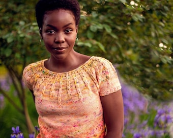 May include: A woman wearing a peach-colored knitted top with a colorful, textured design and a light blue denim skirt. She is standing in a field of purple irises.