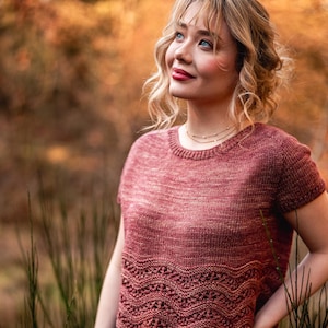 May include: A short-sleeved, reddish-brown knitted top with a scalloped hem. The top is worn with black jeans. The background is blurred, with autumn-coloured foliage and tall green grass.