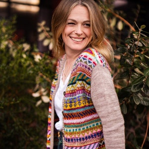 May include: A woman wearing a multi-coloured cardigan with a geometric pattern. The cardigan is knitted with a beige yarn and has a colourful, horizontal stripe pattern. The woman is wearing a white shirt and a plaid skirt.