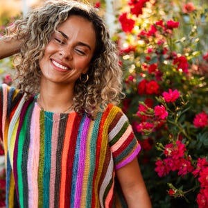 Op de afbeelding: Een gebreide top met korte mouwen en verticale strepen in een levendig kleurenpalet, waaronder felroze, oranje, geel, groen, blauw, paars en bruin. Het kledingstuk heeft een ronde halslijn en een textuur die doet denken aan breiwerk.