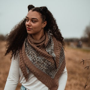 May include: A brown and gray knitted shawl with a textured pattern. The shawl is draped over a long-sleeved light-colored shirt and paired with light blue ripped jeans. The overall aesthetic is earthy and natural, suitable for fall or winter wear.