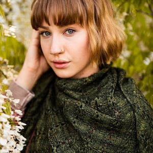 Op de afbeelding: Een vrouw met kort, bruin haar draagt een groene gebreide sjaal met een kantpatroon. Ze staat voor een witte bloeiende struik.
