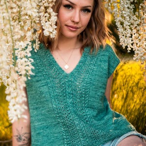 Puede incluir: Una blusa de punto verde turquesa con cuello en V y mangas cortas. La blusa presenta un diseño calado detallado en el centro. La persona lleva pantalones cortos de mezclilla y un collar dorado. Flores blancas en el fondo.