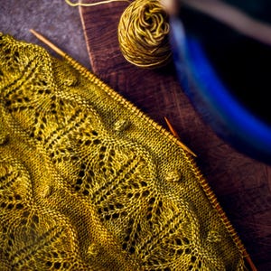 Può includere: Primo piano di un tessuto lavorato a maglia giallo con un motivo traforato. In secondo piano si vede un gomitolo di lana giallo e ferri da maglia.