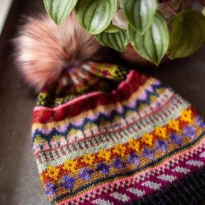 Puede incluir: Un gorro de punto colorido con un pompón en la parte superior. El gorro tiene un patrón geométrico en tonos de rosa, rojo, amarillo, verde, azul y morado.
