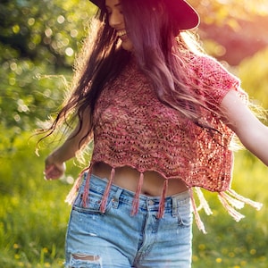 May include: A burgundy fedora hat and a pink crocheted crop top with fringe detail are worn with denim shorts. The top has a detailed openwork pattern. The image is taken outdoors in natural light.