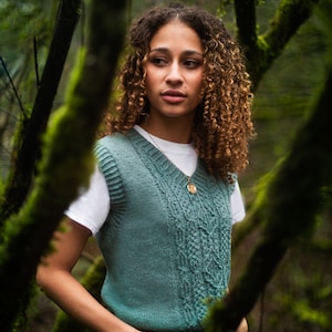 May include: A woman with curly brown hair wearing a teal knitted waistcoat over a white t-shirt and blue jeans. She is standing in a woodland setting with green moss on the trees.