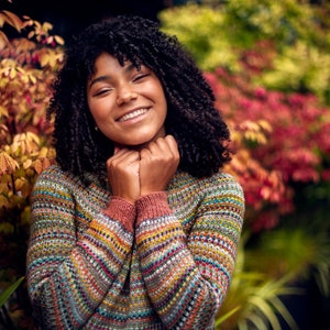 May include: A woman with curly dark hair smiles while wearing a colourful, striped jumper. The jumper has a variety of colours, including red, blue, green, and yellow. The woman is standing in front of a blurred background of autumn foliage.
